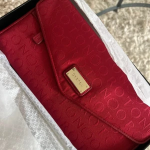 Röd kuvertväska från Oroton - Snygg röd kuvertväska från Oroton med präglad logotyp över hela ytan och guldfärgad metallplatta framtill. Väskan har flera fack, kortplatser och en dragkedja. Materialet är slätt och lyxigt, perfekt för att hålla ordning på dina essentials.