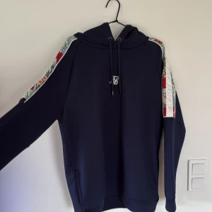 Mörkblå hoodie från SikSilk med blommor - Säljer en mörkblå hoodie från SikSilk med blommiga detaljer och vita ränder längs axlarna. Tröjan har huva med snörning och ett broderat SikSilk-logo på bröstet. Materialet är mjukt och känns som polyester. Perfekt för dig som gillar streetwear med lite extra stil.