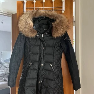 Svart Rockandblue dunjacka med päls - Svart lång dunjacka från Rockandblue med stor fluffig päls runt huvan. Jackan har quiltade detaljer, dragkedjor på fickorna och snygga knappar framtill. Märkespatch på ärmen och figurnära passform. Perfekt för kalla vinterdagar. Observera att där är ett hål i innerfickan, men syns inte utåt.