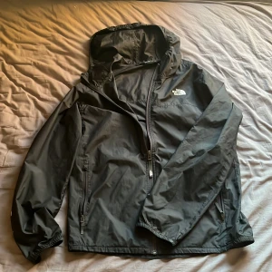 Svart vindjacka från The North Face - Svart vindjacka från The North Face med huva och dragkedja framtill. Jackan har två sidofickor med dragkedja och elastiska muddar vid ärmslut. Perfekt för blåsiga dagar och har en stilren look med logga på bröstet.