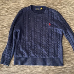 Mörkblå kabelstickad tröja Polo Ralph Lauren - Kabelstickad mörkblå tröja från Polo Ralph Lauren med klassisk rund hals och röd broderad logga på bröstet. Tröjan är långärmad och har ribbade muddar och nederkant. Perfekt för dig som gillar preppy och tidlös stil. (Pris kan diskuteras)