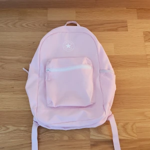 Ljusrosa ryggsäck från Converse - Säljer en ljusrosa ryggsäck från Converse med klassisk logga framtill. Väskan har ett stort huvudfack med ficka för t.ex. papper eller dator, en praktisk ytterficka med dragkedja och meshficka på sidan. Justerbara, vadderade axelremmar för extra komfort. Nypris 400 kr. Perfekt för skolan.🩷📃📐