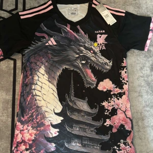Japan Adidas fotbollströja  - Unik svart och rosa Japan fotbollströja från Adidas med stor drake, körsbärsblommor och japanskt tempel. Broderad JFA-logga och rosa Adidas-detaljer. Tillverkad i lätt funktionsmaterial (AEROREADY) med korta ärmar och V-ringning.