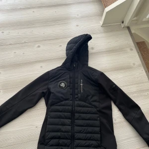 Svart pufferjacka Sail Racing - Svart pufferjacka från Sail Racing med huva och hel dragkedja. Jackan har quiltade partier framtill och på huvan, samt logga på bröstet och ärmen. Snygg och sportig design, perfekt för kyliga dagar.
