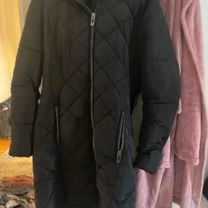 Svart quiltad lång pufferjacka Reserved strl S - Säljer en svart lång pufferjacka med quiltat mönster och dragkedja framtill. Jackan har två stora fickor med dragkedjor och en mysig huva. Perfekt för kalla dagar när du vill hålla dig varm och ändå se stilren ut. Fint skick. 