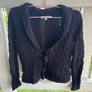 Svart stickad kofta med knyt från First Class - Svart stickad kofta från First Class med snyggt kabelstickat mönster och knyt framtill. Koftan har lång ärm och en lite lösare krage, perfekt att slänga över en topp. Stilren och enkel att matcha till olika outfits.