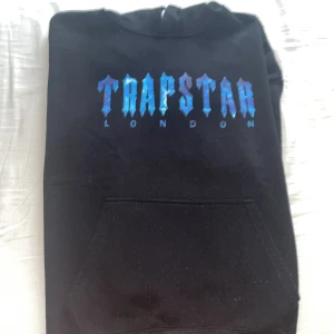 Svart Trapstar hoodie med blått tryck och skön för vintern  - Svart hoodie från Trapstar London med fet blå logga framtill. Klassisk känguruficka och huva. Perfekt för dig som gillar streetwear och vill sticka ut med en snygg detalj i garderoben. Tygen är mjuk och skön