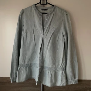 Ljusblå skjorta från KappAhl - Säljer en ljusgrå skjorta från KappAhl med lång ärm och knappar framtill. Skjortan har en rak passform, rund halsringning och volangdetalj nedtill. 