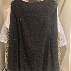 Svart glittrig kortklänning med öppen rygg - Svart glittrig kortklänning med djup öppen rygg och knytband i nacken. Klänningen har en snygg drapering baktill och är tillverkad i ett stretchigt material med metalltrådar som ger extra shimmer. Perfekt för dig som vill sticka ut med en cool look.