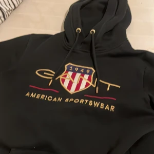 Svart hoodie från GANT  - Svart hoodie från GANT i storlek XS med broderad logga och amerikanskt inspirerat emblem på bröstet. Tröjan har luva med snörning och en stor magficka. Skriv om intresserad pris kan bli mindre om diskuteras 