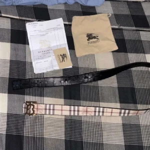 Burberry rutigt bälte med B-spänne - Säljer ett snyggt Burberry-bälte med klassiskt rutigt mönster i beige, svart och rött. Bältet har ett stort metallspänne med Burberrys B-logga och är gjort i skinn. Perfekt accessoar för att lyfta din outfit med en lyxig touch.