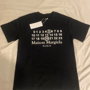 Maison Margiela svart t-shirt - Svart t-shirt från Maison Margiela med ikonisk vit print framtill med siffror och logga. Klassisk rund hals och korta ärmar. Materialet är mjuk bomull och passformen är normal. Perfekt för dig som gillar stilrena designer med en twist. 
