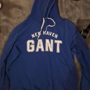 Blå hoodie från GANT med tryck - Snygg blå hoodie från GANT med vit text 'NEW HAVEN GANT' på bröstet. Tröjan har huva med vita snören och är långärmad. Perfekt för dig som gillar klassisk college-stil och vill ha något bekvämt och stilrent.