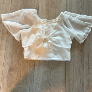 Vit blus med knyt från Lindex - Supersöt vit blus från Lindex med kort croppad passform och vida, transparenta ärmar. Toppen har en snygg knytning framtill och är gjord i ett lätt, luftigt material som passar perfekt till varma dagar. Perfekt för dig som gillar fina blusar och trendig stil.💕
