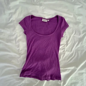 Lila ribbad topp från Kappahl - En lila ribbad topp från Kappahl med korta ärmar och rund, djup urringning. Toppens material är stretchigt och sitter snyggt mot kroppen. Perfekt att styla med jeans eller kjol för en färgstark look.