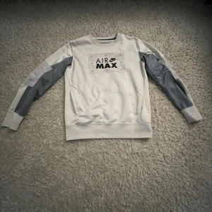 Nike Air Max sweatshirt i grått - Nike Air Max sweatshirt i ljusgrått med mörkgrå paneler på ärmarna och tryckt logga framtill. Klassisk crewneck-modell med ribbade muddar och mjukt material. Perfekt för en sportig och avslappnad stil.