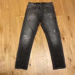 Svarta Jack and Jones jeans med slitningar - Snygga svarta Jack and Jones jeans med coola slitningar på framsidan. De har en klassisk femficksdesign och en knappgylf. Jeansen är i storlek W28 men sitter bra på 30 också