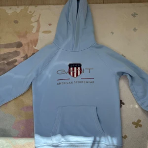 Ljusblå hoodie från Gant - Snygg ljusblå hoodie från Gant med klassisk logga och texten 'American Sportswear' framtill. Tröjan har huva, magficka och långa ärmar. Perfekt för en chill och sportig look. Materialet känns mjukt och skönt mot huden.