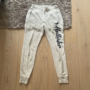 Gråa mjukisbyxor från Hollister - Snygga beiga mjukisbyxor från Hollister med mörkblå logga längs benet. Byxorna har resår och snörning i midjan, ribbade muddar vid bensluten och två fickor framtill. Perfekta för chill dagar eller när du vill vara bekväm.