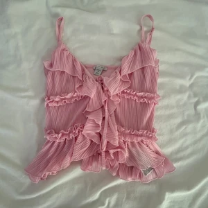 Rosa volangtopp från Amisi XS - Supersöt rosa linnetopp från Amisi i storlek XS. Toppen har tunna axelband, volanger och plisserat tyg med knytning framtill. Perfekt för dig som gillar romantisk och feminin stil. Luftig och flowy känsla, passar grymt till jeans eller kjol.