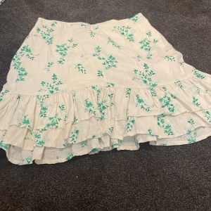 Vit blommig volangkjol från Lindex - Supersöt vit kjol från Lindex med grönt blommönster och flera volanger. Kjolen har en kort modell och är gjord i mjuk bomull, perfekt för sommarens alla äventyr. Elastisk midja för skön passform och flowig känsla.