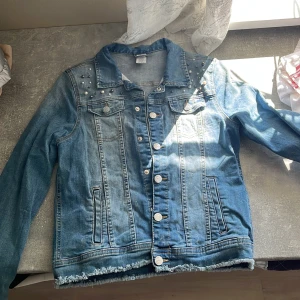 Jeansjacka med pärlor från Lindex - Blå jeansjacka från Lindex med snygga pärldetaljer på axlarna och fransad nederkant. Klassisk modell med knappar framtill, två bröstfickor och långa ärmar. Perfekt för dig som vill ha en cool och unik denimlook.