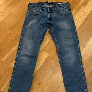 Replay blå jeans slim fit - Snygga blå jeans från Replay med klassisk femficksdesign och coola slitningar. Modellen har smal passform och raka ben, med tydliga kontrastsömmar och Replay-logga på bakfickan. Perfekta för dig som gillar en stilren och modern look.