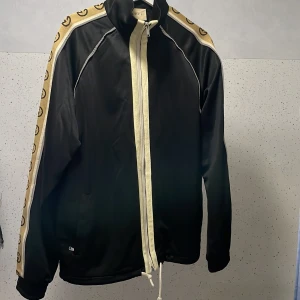 Svart Gucci vindjacka med logoband - Gucci vindjacka i svart med krämfärgade detaljer och dragkedja framtill. Ärmarna har breda logoband i beige med bruna GG-symboler. Jackan har hög krage, snörning nertill och sportig vibe. Perfekt för dig som vill sticka ut med lyxig streetstyle.