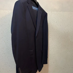 Mörkblå wool stretch kostym, regular fitDressmann slim fit 48 - Snygg smörkblå  helt sätt  från Dressmann i slim fit-modell, storlek 48. Kavajen har klassisk design med smala slag, knappar vid ärmslut och innerfoder i grått med röda detaljer. Perfekt för dig som vill ha en stilren och modern look.