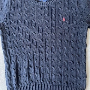 Mörkblå stickad tröja Polo Ralph Lauren - Kabelstickad mörkblå tröja från Polo Ralph Lauren med normal långa ärmar och klassisk rund hals. Tröjan har en röd broderad logga på bröstet och ribbade muddar nedtill. Perfekt för dig som gillar preppy och stilren look.