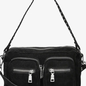 Svart mocka axelväska med dragkedjor - Cool svart axelväska i mocka med två stora framfickor och silvriga dragkedjor. Väskan har justerbar axelrem med snygga detaljer och flera fack för att hålla ordning på dina grejer. Perfekt för dig som vill ha en stilren men edgy look.