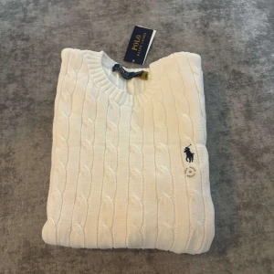 Vit stickad tröja från Polo Ralph Lauren - Vit kabelstickad tröja från Polo Ralph Lauren med klassisk rund hals och broderad marinblå logga på bröstet. Tröjan har långa ärmar och ribbade muddar vid hals, ärmslut och nederkant. Perfekt för dig som gillar clean och stilren look.