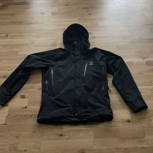 Svart regnjacka från Haglöfs - Snygg svart regnjacka från Haglöfs med huva och flera praktiska dragkedjor på bröstet och sidorna. Jackan har en clean look, långärmad design och är tillverkad i slitstark polyester som står emot regn och vind. Perfekt för dig som gillar att vara ute oavsett väder. Knappast använd och storlek M Usa