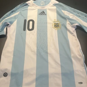 Argentina Messi 10 Adidas fotbollströja -  Jag behöver sälja snabbt! Säljer en officiell Argentina fotbollströja från Adidas med Messis namn och nummer 10. Tröjan har klassiska ljusblå och vita ränder, broderat AFA-emblem och Adidas-logga. Tillverkad i lätt och ventilerande Clima365-material, kortärmad och perfekt för fotbollsfans.
