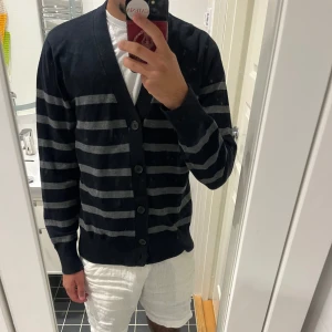 Svart och grå randig kofta Tommy Hilfiger - Snygg svart kofta från Tommy Hilfiger med gråa horisontella ränder. Koftan har v-ringning, knäppning framtill med svarta knappar och långa ärmar. Tillverkad i mjuk bomull, perfekt för lager-på-lager och chill stil.