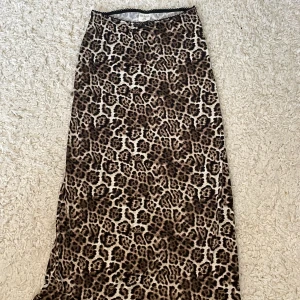 Leopardmönstrad långkjol från Stories XS - Säljer en snygg långkjol från Stories i storlek XS med leopardmönster i bruna och beige toner. Kjolen har en liten svart rosett framtill och dekorativ spetskant upptill. Perfekt för dig som vill sticka ut med djurmönster.