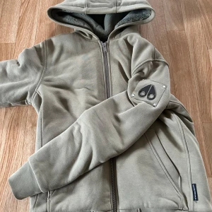  Grön Moose Knuckles bunny jacka  - Snygg beige/grön hoodie/jacka från Moose Knuckles med dragkedja och huva. Insidan är fodrad med mjuk fleece och quiltat svart innerfoder. Ärmen har den ikoniska logotypen i metall. Perfekt för kyliga dagar och riktigt skön att bära. Inga skador och använd 4 gånger. Kan mötas upp. Accepterar byten endast jackor.