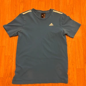 Blå Adidas t-shirt med vita detaljer - Snygg blå t-shirt från Adidas med vita ränder på axlarna och klassisk Adidas-logga på bröstet. T-shirten har rund hals och korta ärmar, perfekt för en sportig look. Materialet känns mjukt och skönt mot huden.