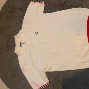 Vit t-shirt med dragkedja - Vit t-shirt med kort ärm, diskret ribbad struktur och klassisk krage. Dragkedja framtill och broderad logga på bröstet. Röd och svart randig detalj på ärmslut och nederkant. Perfekt för en clean och sportig look.