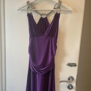 Lila vintage långklänning med kedjedetaljer  - Snygg lila långklänning med v-ringning och draperad midja. Klänningen har unika axelband i silverfärgad kedja som korsas i ryggen och ger en cool statement-look. Ger dig en elegant, snygg och modern stil. Passar S-M