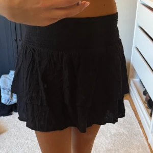 Svart volangkjol med resårmidja - Supersöt svart kortkjol med volanger och bred smockad resår i midjan. 