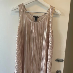 Champagnefärgad plisserad klänning  - Snygg elegant och modern look, ärmlös klänning från H&M i skimrande champagnefärg. Klänningen är plisserad med ett lätt glansigt tyg och har en rak, luftig passform. Den skulle passa jättebra med ett stort bälte dessutom! Pris går att diskuteras 