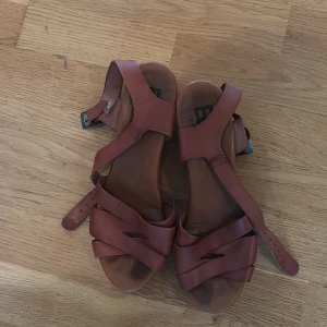 Bruna sandaler från Eden of Sweden - Snygga bruna sandaler med flera breda remmar i skinn och justerbart spänne vid ankeln. Modellen har öppen tå och platt sula, perfekt för dig som gillar en chill och stilren look. Passar dig som vill ha något enkelt men ändå trendigt till sommaren.