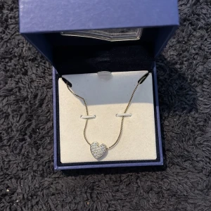 Swarovski halsband med glittrigt hjärta - Supersnyggt halsband från Swarovski med ett glittrigt hjärthänge i silverfärg. Kedjan är tunn och elegant, och hjärtat är täckt av små gnistrande stenar som verkligen poppar. Perfekt för dig som vill ha något extra till din look.