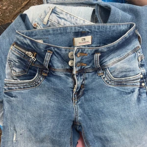 Blå jeans från LTB, W26 L32 - Säljer ett par snygga blå jeans från LTB med coola slitningar och kontrastsömmar. Jeansen har dubbla knappar i midjan, dragkedjedetalj vid fickan och klassisk femficksdesign. Perfekt för dig som gillar en avslappnad men trendig look. Dom har skvätt av målarfärg på sig som där en detalj (fanns där när jag beställde dom)