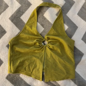 Glittrig gul halterneck topp - Snygg glittrig gul halterneck topp med ringdetalj framtill och öppen slits nertill. Toppen är croppad och har en ärmlös design som går runt nacken. Perfekt för dig som vill sticka ut med en cool och trendig look. Toppen är aldrig använd.