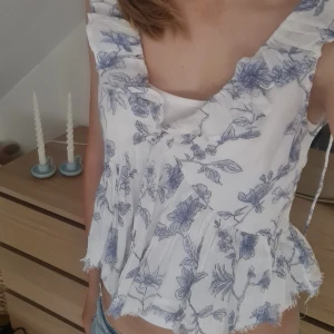 Blommig volangblus från Gina Tricot XS - Supersöt blus från Gina Tricot i vitt med blått blommönster. Modellen har volangdetaljer längs axelbanden och nederkanten, omlottkänsla och lätt rynkade partier. Luftig och somrig med romantisk vibe, perfekt till jeans eller kjol. Är aldrig använd och har prislappen kvar. Nypris 299