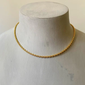 Guldfärgat tvinnat halsband - Snyggt halsband i guldfärgad metall med tvinnad kedja. Kedjan har en klassisk karbinhake och justerbar längd för perfekt passform. Enkel men ändå stilren design som lyfter vilken outfit som helst. 43 cm