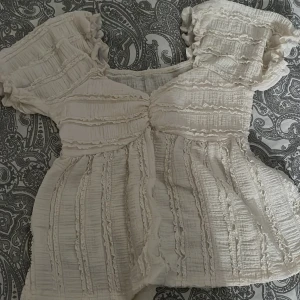 Romantisk volangblus i ljus beige - Supersöt blus i ljus beige med volangdetaljer och rynkade partier. Blusen har korta puffärmar och v-ringning med rynk framtill. Perfekt för dig som gillar en romantisk och bohemisk stil.