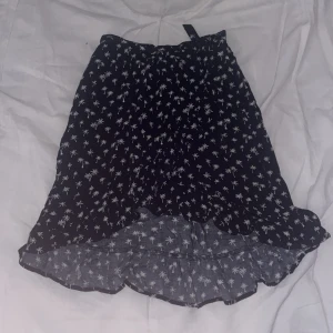 Svart kjol med palmmönster och volang - Svart kortkjol med vita palmmönster och volangkant längst ner. Kjolen har en lätt och flowy känsla, perfekt för varma dagar. Resår i midjan och liten knyt-detalj upptill. Snygg till sneakers eller sandaler.